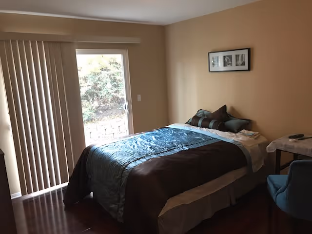 A cozy bedroom with a made bed topped with decorative pillows, a sliding glass door with vertical blinds, and a small side table and chair.