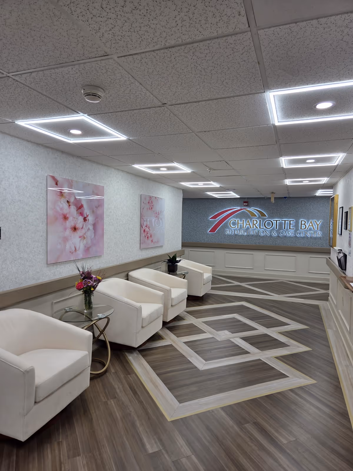 A well-lit waiting area with four white armchairs arranged along a wall. There are two small glass side tables, one with a vase of flowers and the other with a small plant. The floor has a decorative geometric pattern in wood tones. The wall features two floral artworks and a large illuminated sign that reads 'Charlotte Bay Rehabilitation & Care Center'. The ceiling has recessed lighting with square light fixtures.
