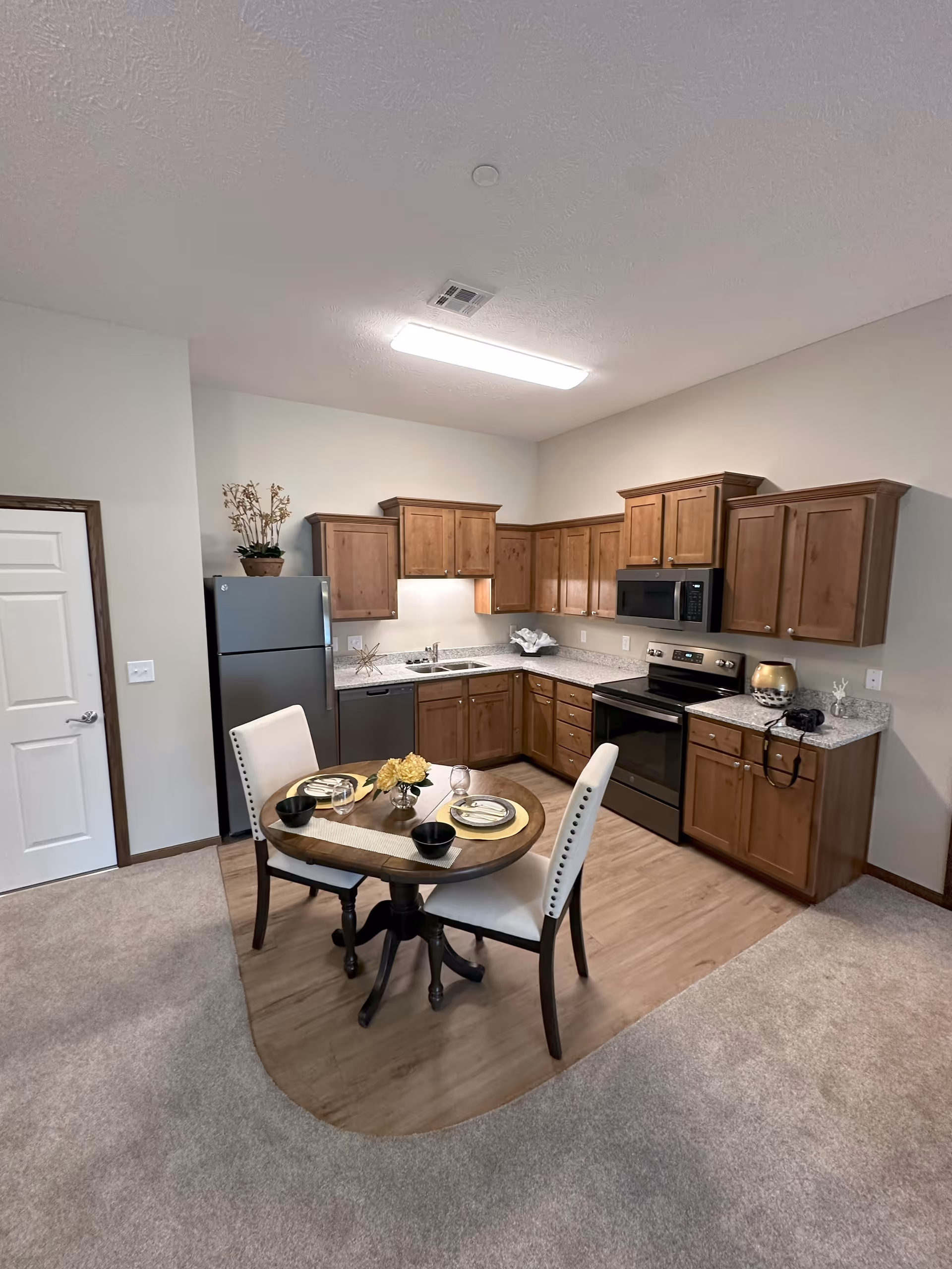 A small kitchen area with wooden cabinets, a stainless steel refrigerator, dishwasher, stove, and microwave. In front of the kitchen is a round wooden dining table set for two with white cushioned chairs. The floor transitions from carpet to wood in the kitchen area. The walls are light-colored and there is a ceiling light fixture.