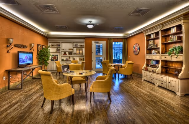 A cozy common area with wooden flooring and warm orange walls. The room features several yellow upholstered chairs arranged around small round tables. There is a large wooden bookshelf filled with books and decorative items on the right side, and a flat-screen TV mounted on a wooden stand on the left wall. A door and window at the back let in natural light, and the ceiling has recessed lighting and vents.