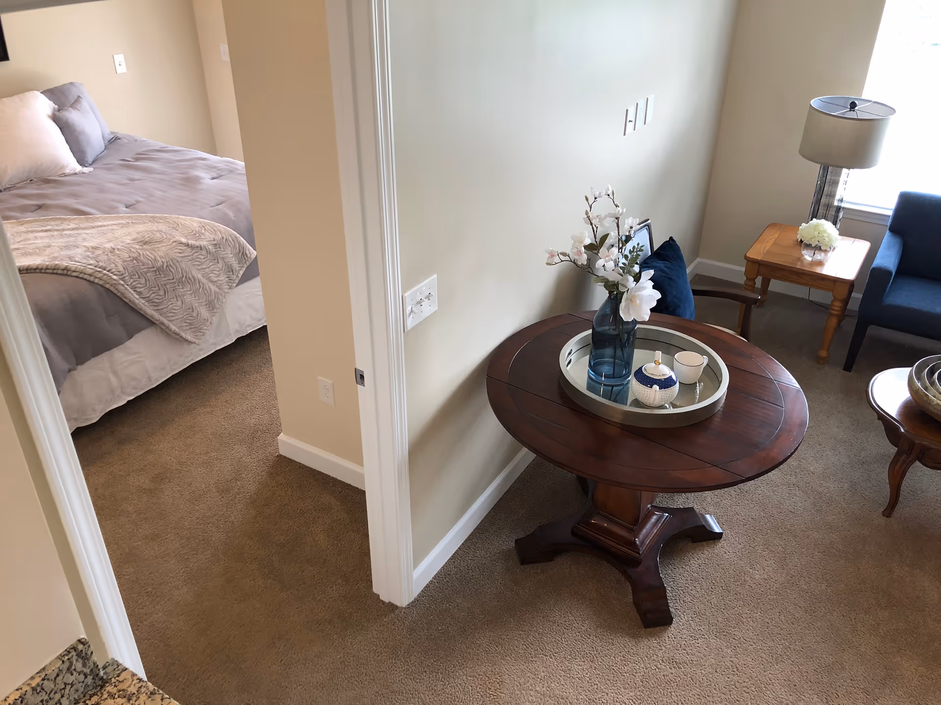 View of a cozy interior space showing a round wooden table with a tray holding a vase with flowers and a small decorative container. To the left, there is a bedroom with a made bed featuring gray bedding and a beige blanket. To the right, part of a living area is visible with a wooden side table, a lamp, a small flower arrangement, and a blue armchair near a window.