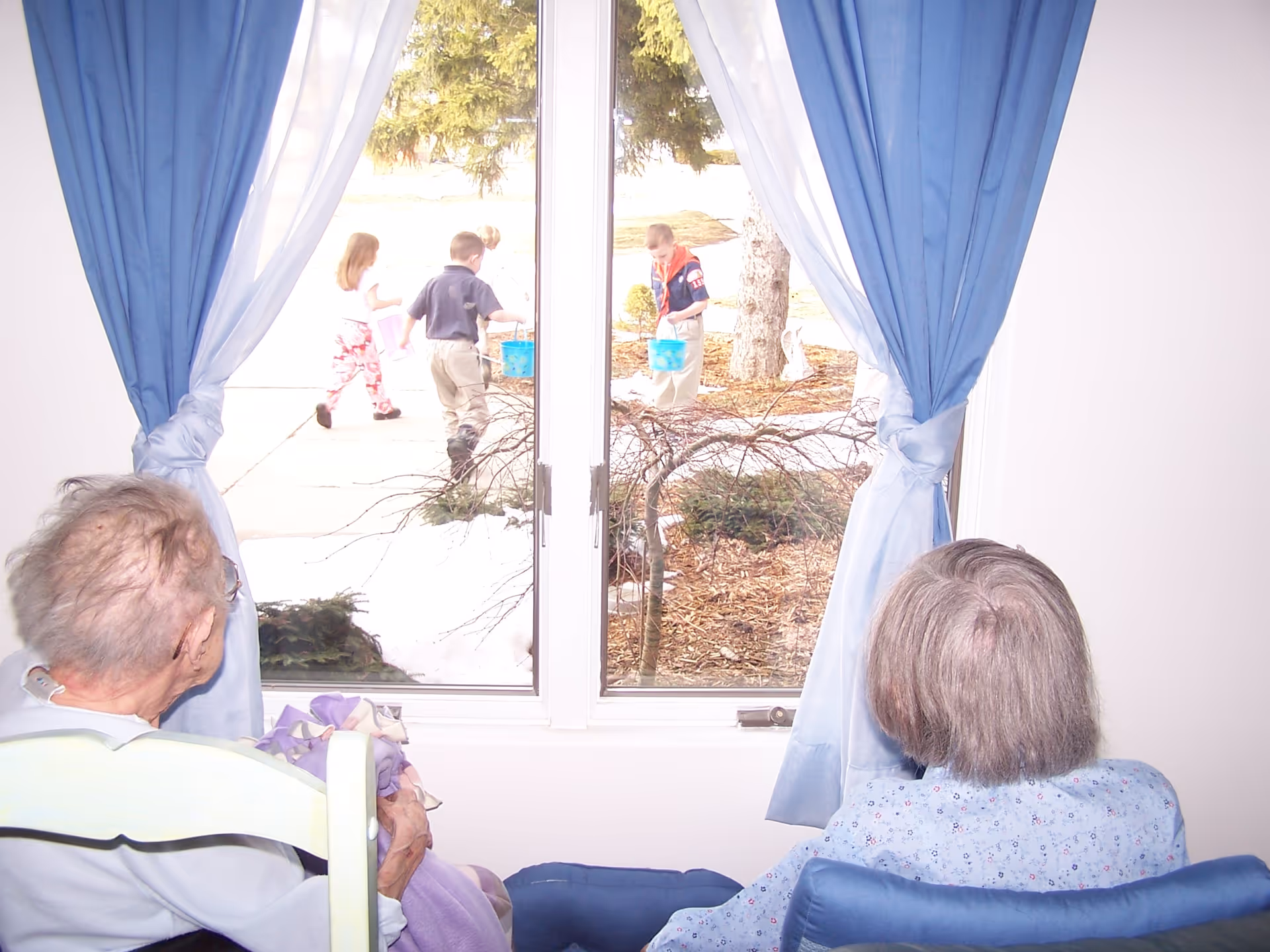 Two elderly residents sit by a window with blue curtains watching children playing outside.