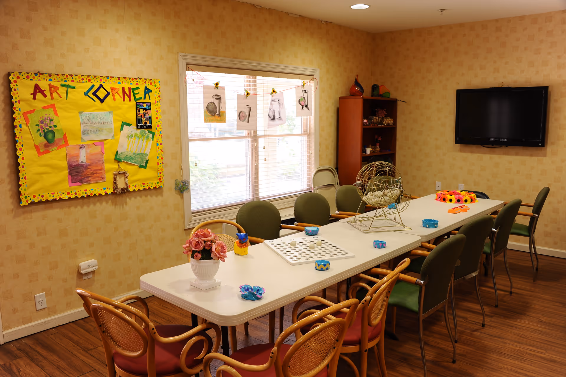 Community activity room with long tables and chairs, a bingo cage, colorful decorations and an 'Art Corner' bulletin board.