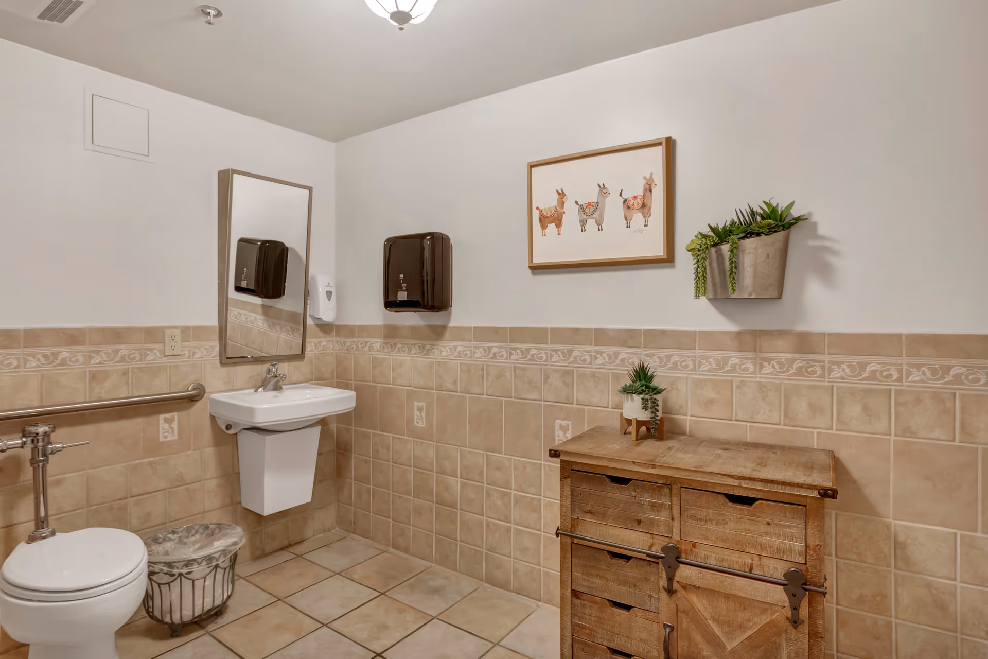 A clean bathroom with beige tiled walls and floor. The bathroom features a white toilet with a metal grab bar, a white wall-mounted sink with a mirror above it, a black paper towel dispenser, and a white soap dispenser. There is a wooden cabinet with drawers on the right side, decorated with two small potted plants. Above the cabinet, a framed picture of four llamas hangs on the wall.