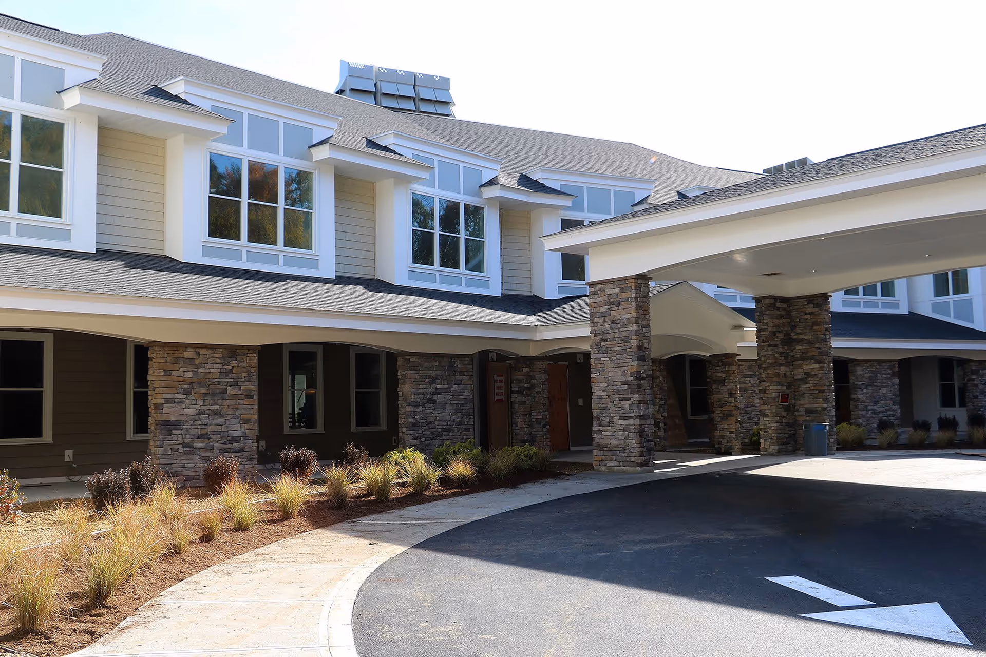 Exterior view of a senior living facility building with stone pillars supporting a covered entrance. The building has multiple large windows and a landscaped area with plants along the sidewalk.