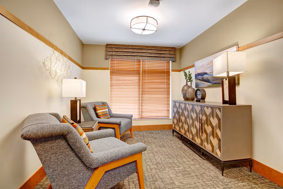 A cozy sitting area with two gray upholstered armchairs featuring wooden legs and colorful striped pillows. Between the chairs is a small wooden side table with a lamp. Opposite the chairs is a decorative wooden cabinet with a geometric pattern, topped with two vases, a clock, and a lamp. A large window with wooden blinds and a valance lets in natural light. The walls are painted beige with a wooden trim, and the floor is carpeted in a patterned design.