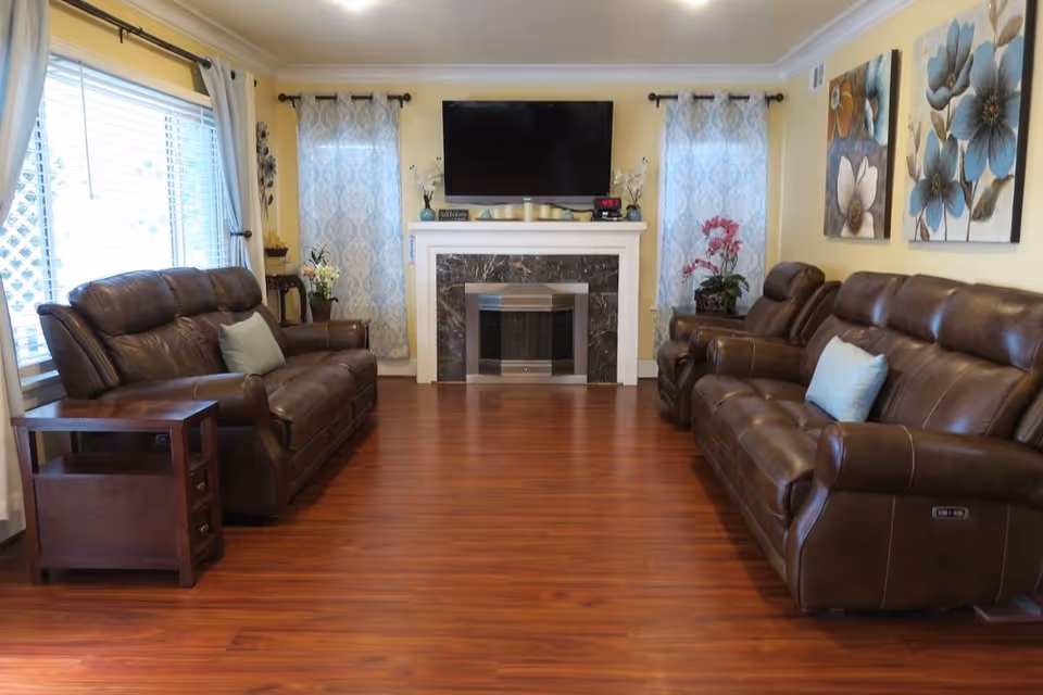 A cozy living room with two brown leather sofas facing each other across a wooden floor. A fireplace with a black marble surround and a mounted flat-screen TV above it is centered on the far wall. Two windows with patterned curtains flank the fireplace, and there are decorative flowers and artwork on the walls.