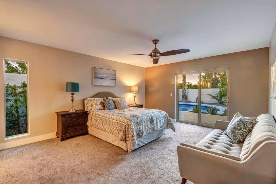 A bright master bedroom with a bed, nightstands and lamps, a tufted sofa, ceiling fan, and sliding glass doors opening to an outdoor pool.