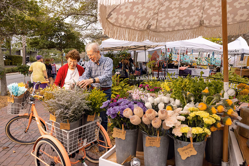 Two older adults browse buckets of flowers at a busy outdoor market stall under umbrellas.
