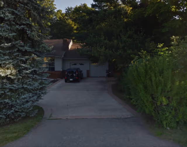 Driveway leading to a single-story house with a garage and a black vehicle parked in front. The house is surrounded by tall trees and bushes.