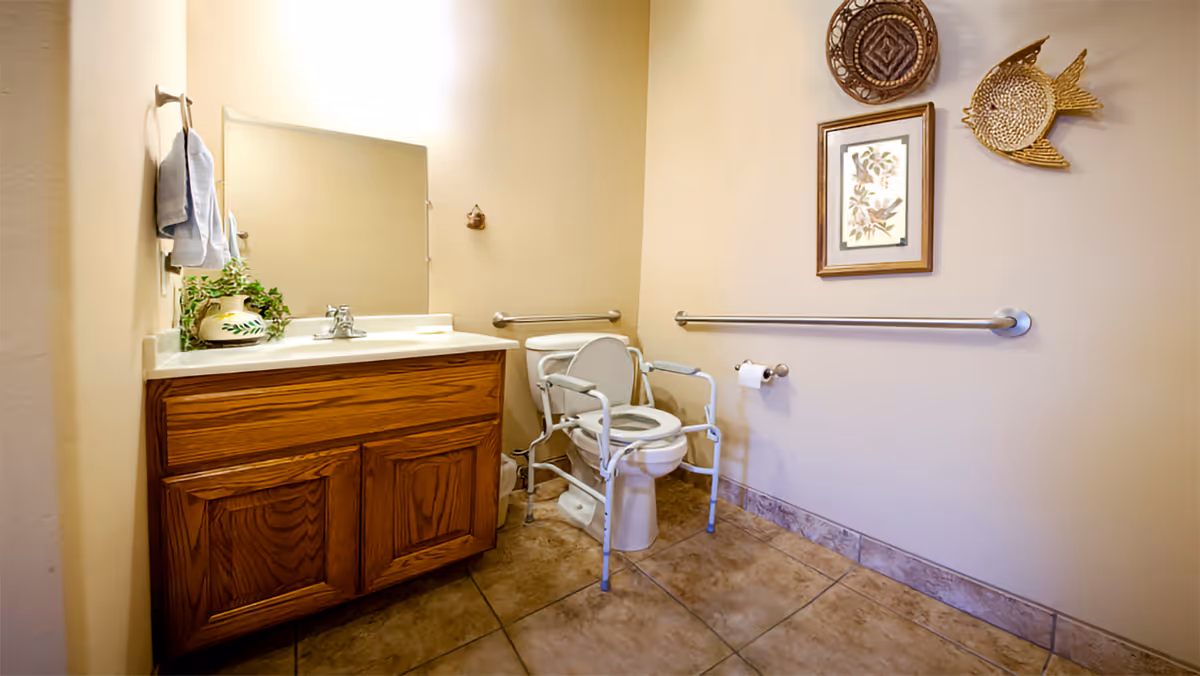 A bathroom with beige walls and tiled floor featuring a wooden vanity with a sink and mirror above it. There is a toilet with a white safety frame around it, a grab bar on the wall, a toilet paper holder, and decorative wall hangings including a framed picture and woven baskets.