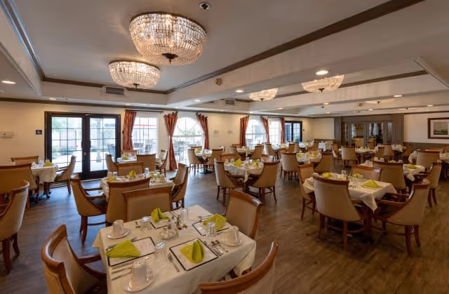 A spacious dining room with multiple tables set for a meal, each table covered with white tablecloths, green folded napkins, cups, and silverware. The room features wooden flooring, large windows with red curtains, and elegant chandeliers hanging from the ceiling.
