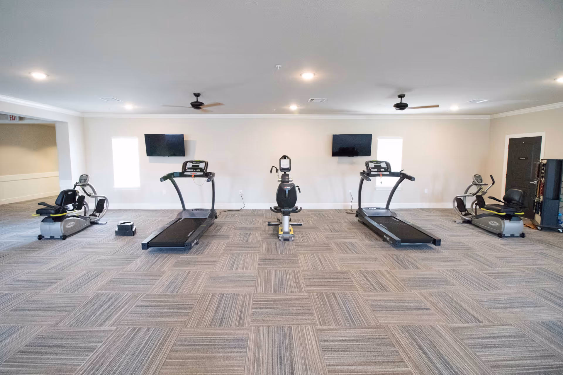 A spacious fitness room with two treadmills, two recumbent exercise bikes, and one upright exercise bike. The room has light-colored walls, two small windows, two ceiling fans, and two wall-mounted televisions. The floor is covered with patterned carpet tiles.