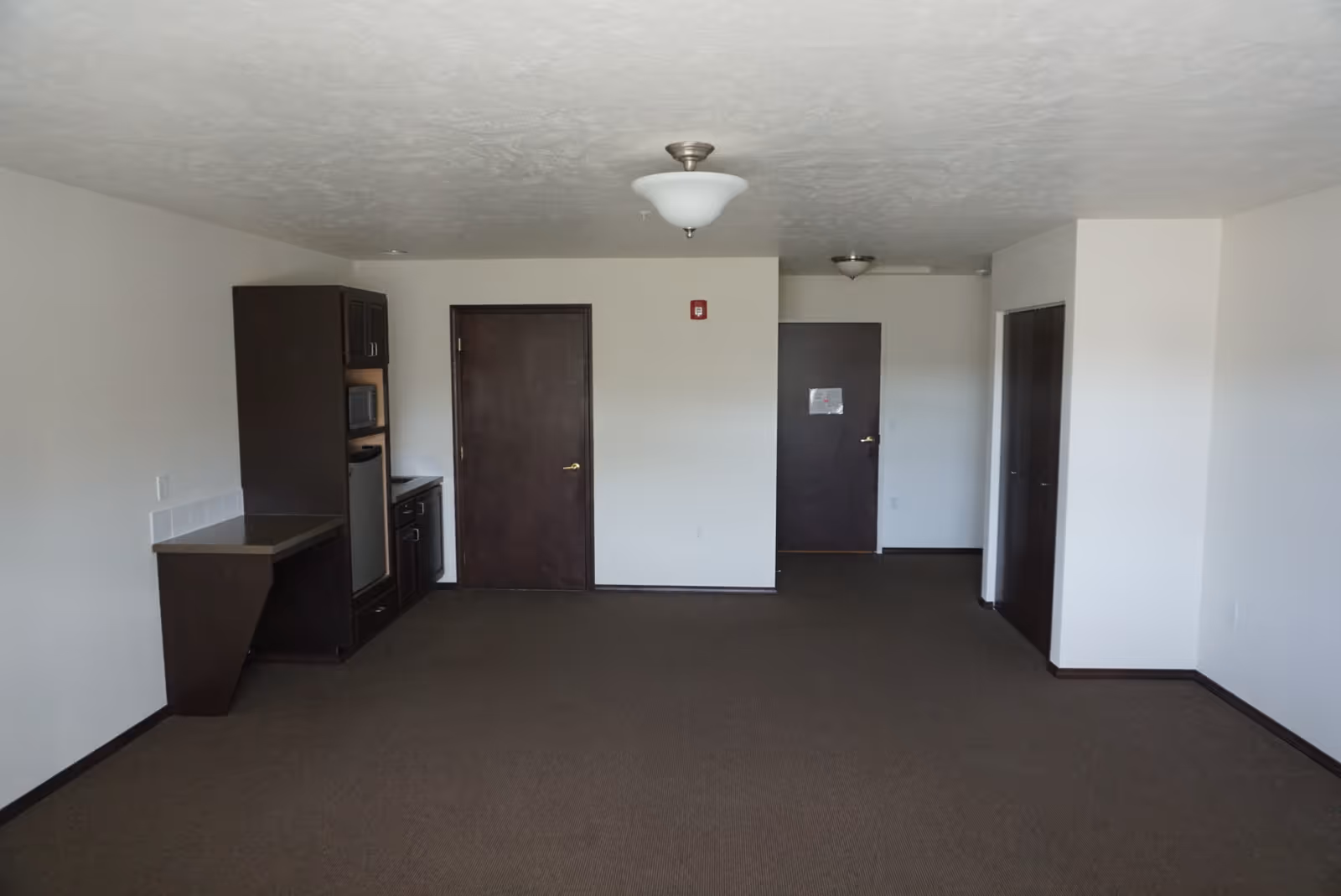 Empty interior room with brown carpet and white walls, featuring dark wood doors and cabinetry including a small kitchenette area with a microwave and mini fridge. Ceiling lights are visible and there is a fire alarm on the wall.