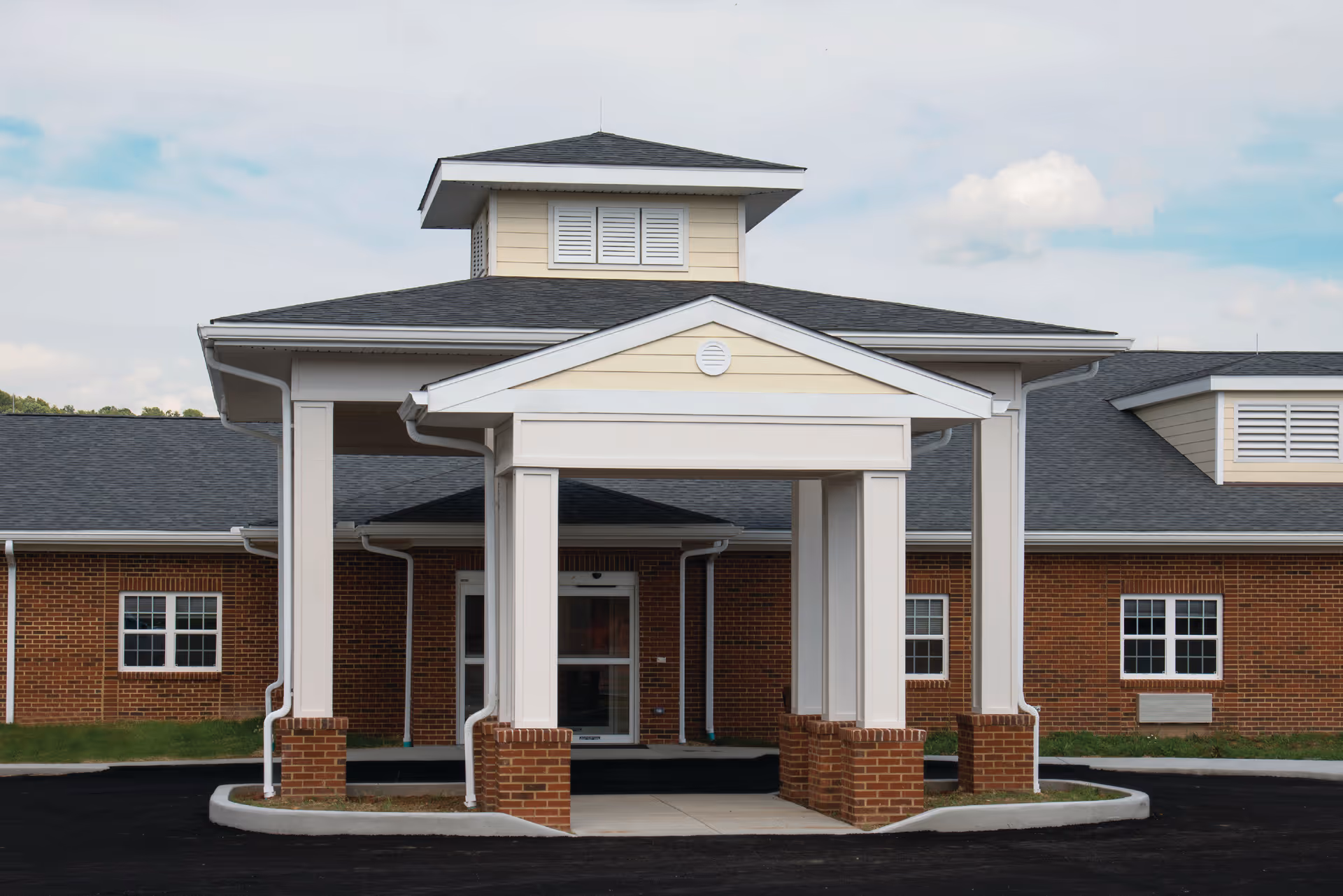 Front exterior view of Maplewood Healthcare Center showing a brick building with a covered entrance supported by white columns and a peaked roof. The sky is partly cloudy.