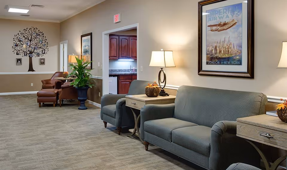 Seating area in a senior living facility with sofas, armchairs, side tables and lamps, wall art, a plant, and a doorway to a small kitchenette.