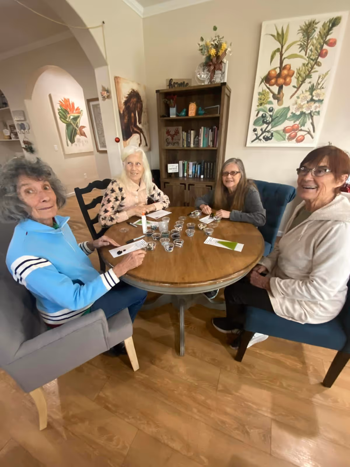Four elderly women sitting around a round wooden table in a cozy room, engaging in an activity with small cups and papers. The room has wooden flooring, a bookshelf with books and decorations, and botanical and horse-themed artwork on the walls.