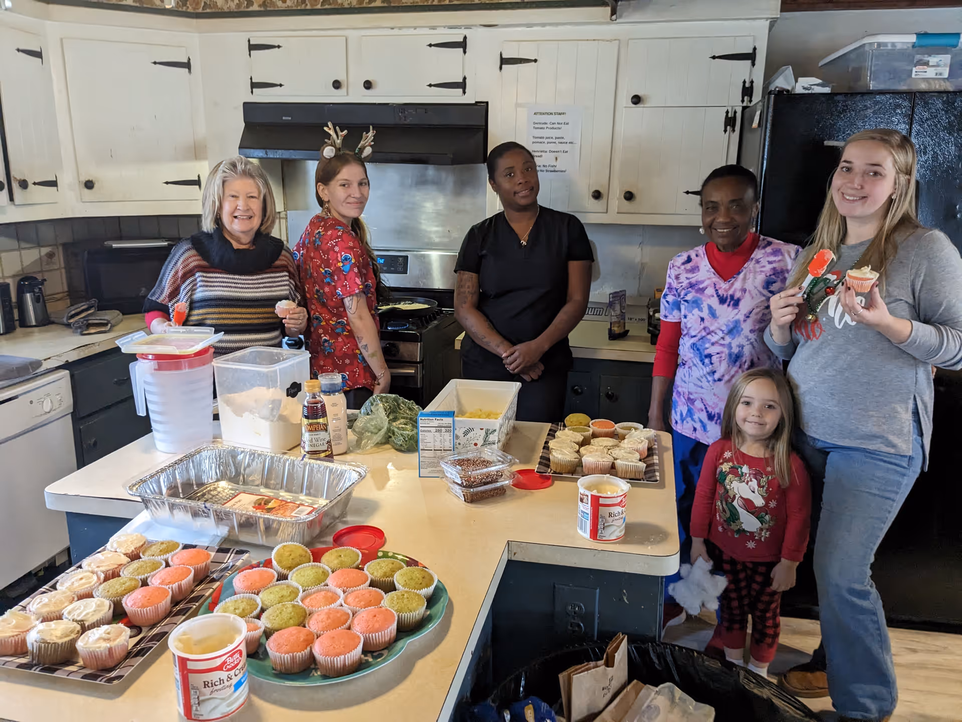 Five women and one young girl standing in a kitchen around a counter with trays of colorful cupcakes and baking supplies. The kitchen has white cabinets, a black refrigerator, and a stove with a hood. The group is smiling and appears to be enjoying a baking activity together.