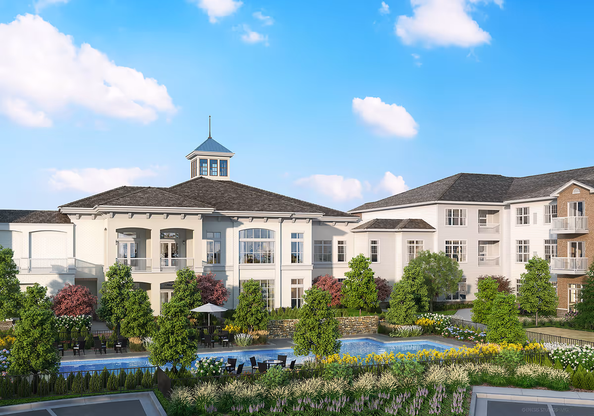 Exterior view of The Sheridan at Eastside senior living facility showing a large, multi-story building with white and beige siding, multiple windows, balconies, and a central cupola. The foreground features a landscaped garden with trees, flowering plants, and a rectangular swimming pool surrounded by patio chairs and tables under umbrellas. The sky is clear with a few clouds.