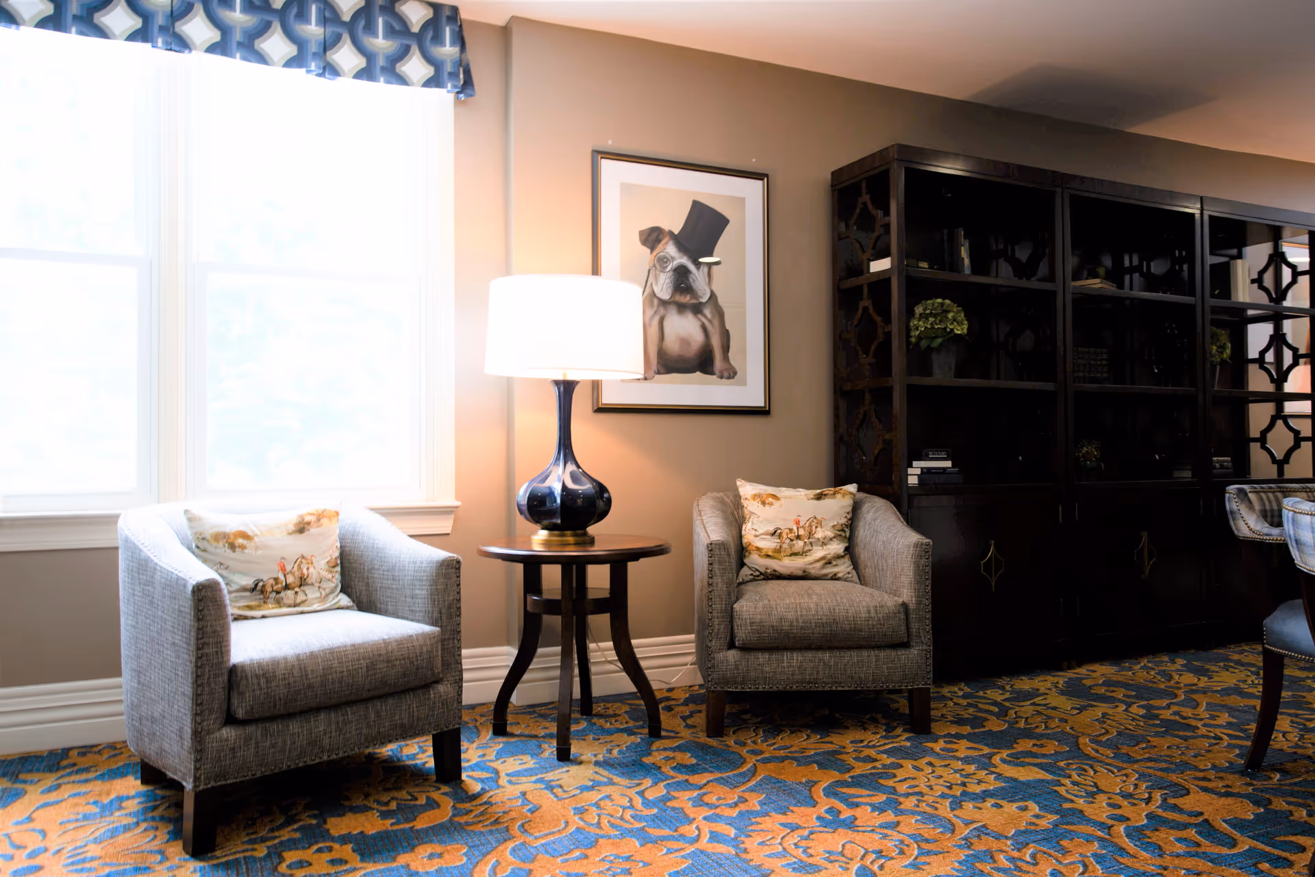 A cozy sitting area with two gray armchairs, each with a decorative pillow featuring a horse design. Between the chairs is a small round wooden table with a blue glass lamp. Behind the table is a framed picture of a bulldog wearing a top hat. To the right is a large dark wooden cabinet with shelves containing books and plants. The floor is covered with a blue and orange patterned carpet, and a window with a blue and white valance lets in natural light.