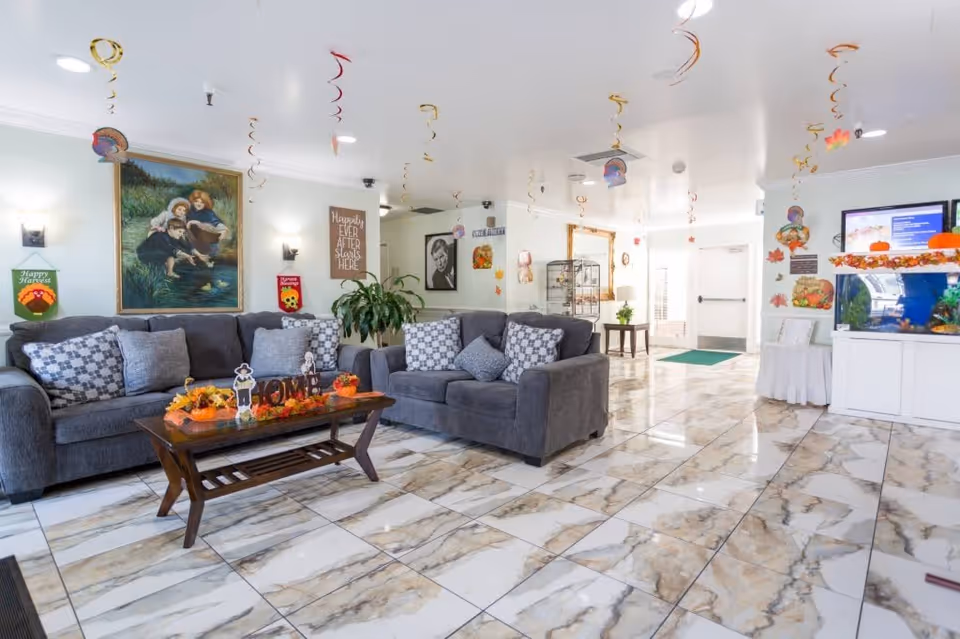 A bright and spacious living room area with two gray sofas adorned with patterned and solid pillows. A wooden coffee table in front of the sofas is decorated with autumn-themed items including small pumpkins and fall leaves. The floor is covered with large, glossy marble tiles. The ceiling has hanging spiral decorations with small turkey images, suggesting a Thanksgiving or fall theme. The walls feature framed artwork and signs, one of which reads 'Hospitality Ever After Starts Here.' There is a white cabinet with an aquarium on the right side and a doorway leading to another area in the background.