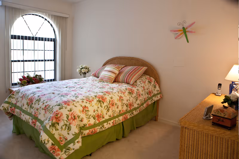 A cozy bedroom with a floral bedspread and striped pillows on a wicker bed frame. There is a wicker dresser with a lamp, a phone, and decorative items. A large arched window with vertical blinds lets in natural light. A colorful dragonfly decoration is mounted on the wall.