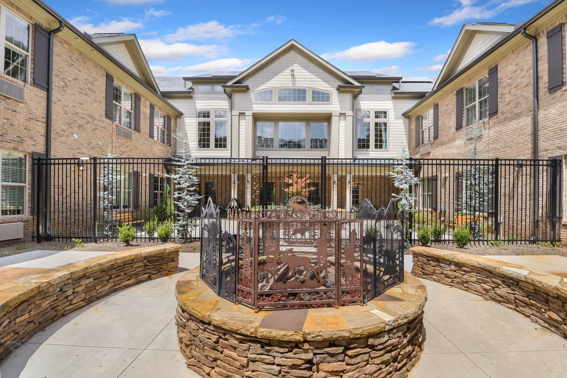 Outdoor courtyard area of a senior living facility with a circular stone fire pit surrounded by a decorative metal screen featuring animal and mountain designs. The courtyard is enclosed by a black metal fence and flanked by two brick buildings with multiple windows. The sky is blue with some clouds.