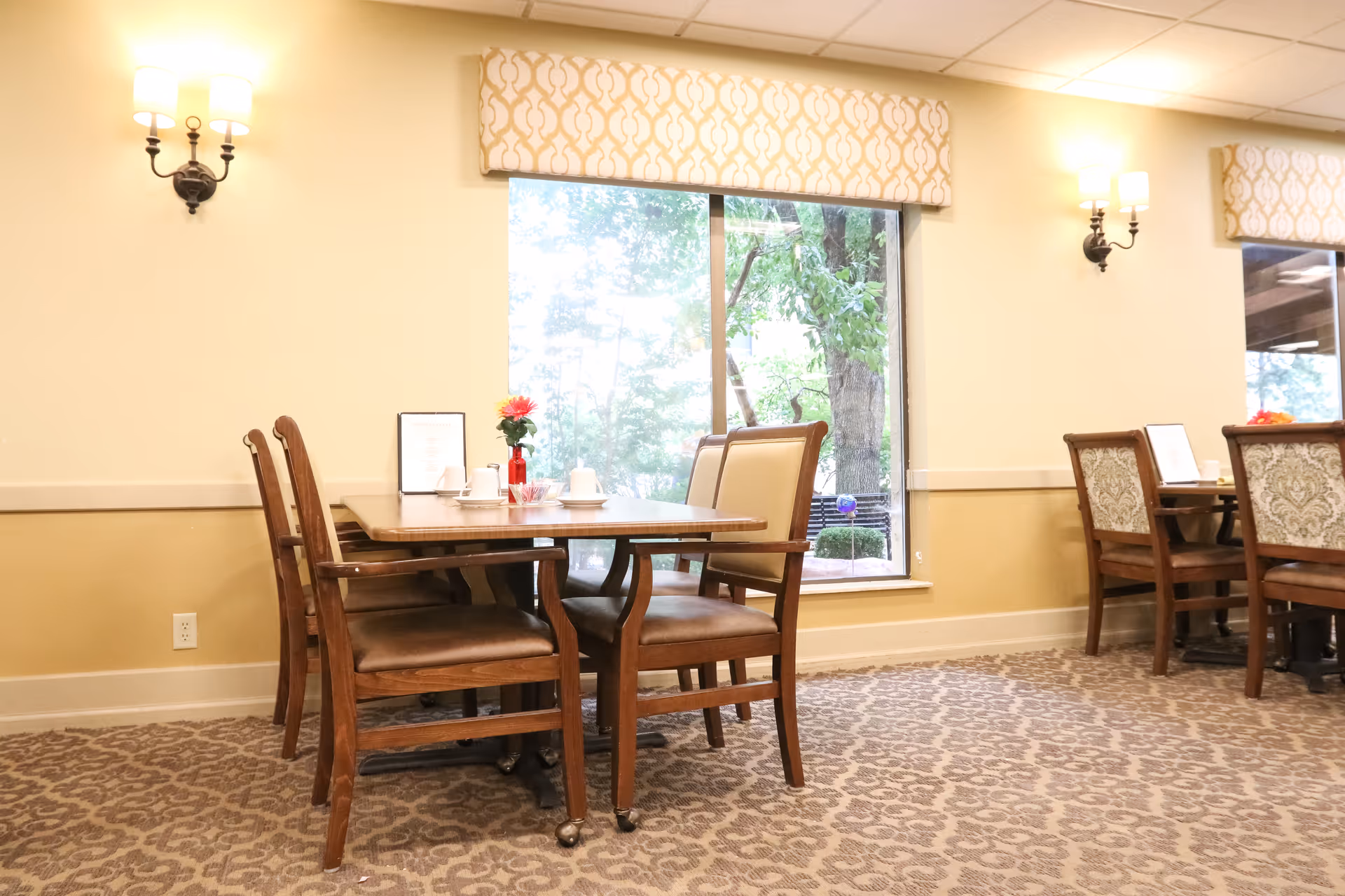 Dining room with wooden tables and chairs, a patterned carpet, wall sconces, and a large window showing greenery.
