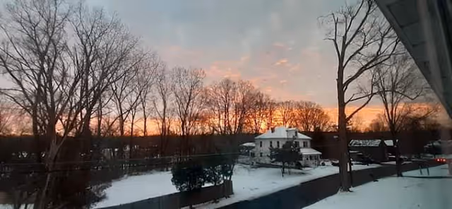 Snow-covered yard and fence with leafless trees and a white house beneath a colorful sunrise sky.