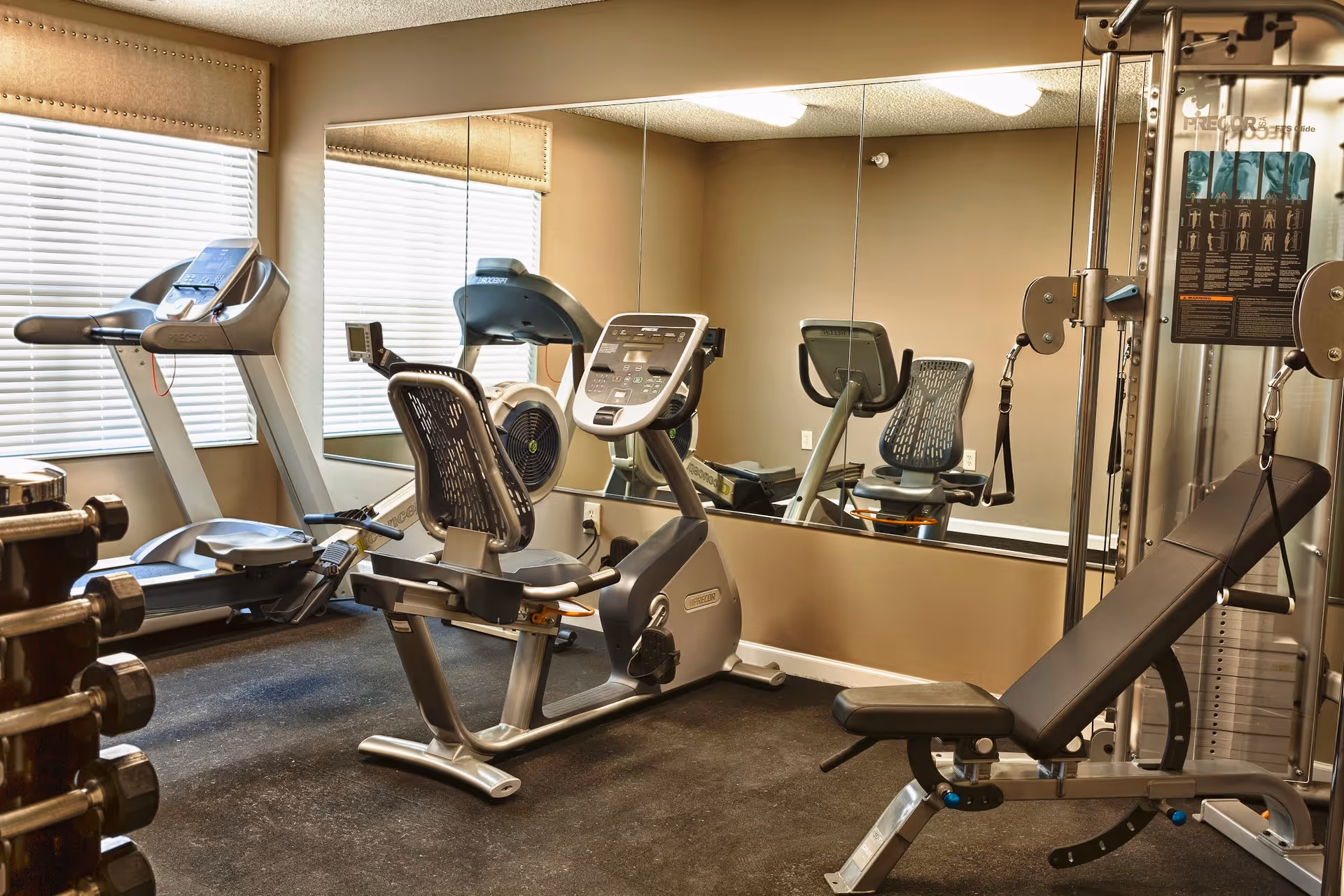 A small fitness room with exercise equipment including a treadmill, a recumbent stationary bike, a weight bench, and a multi-function weight machine. There is a large mirror on one wall and windows with blinds on another wall.