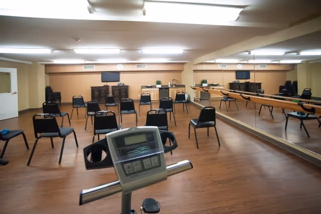 A spacious room with wooden flooring and beige walls, featuring several black chairs arranged in rows facing a wall with a TV and cabinets. A large mirror covers one side of the room, reflecting the chairs and a wooden barre. In the foreground, part of an exercise bike's console is visible.
