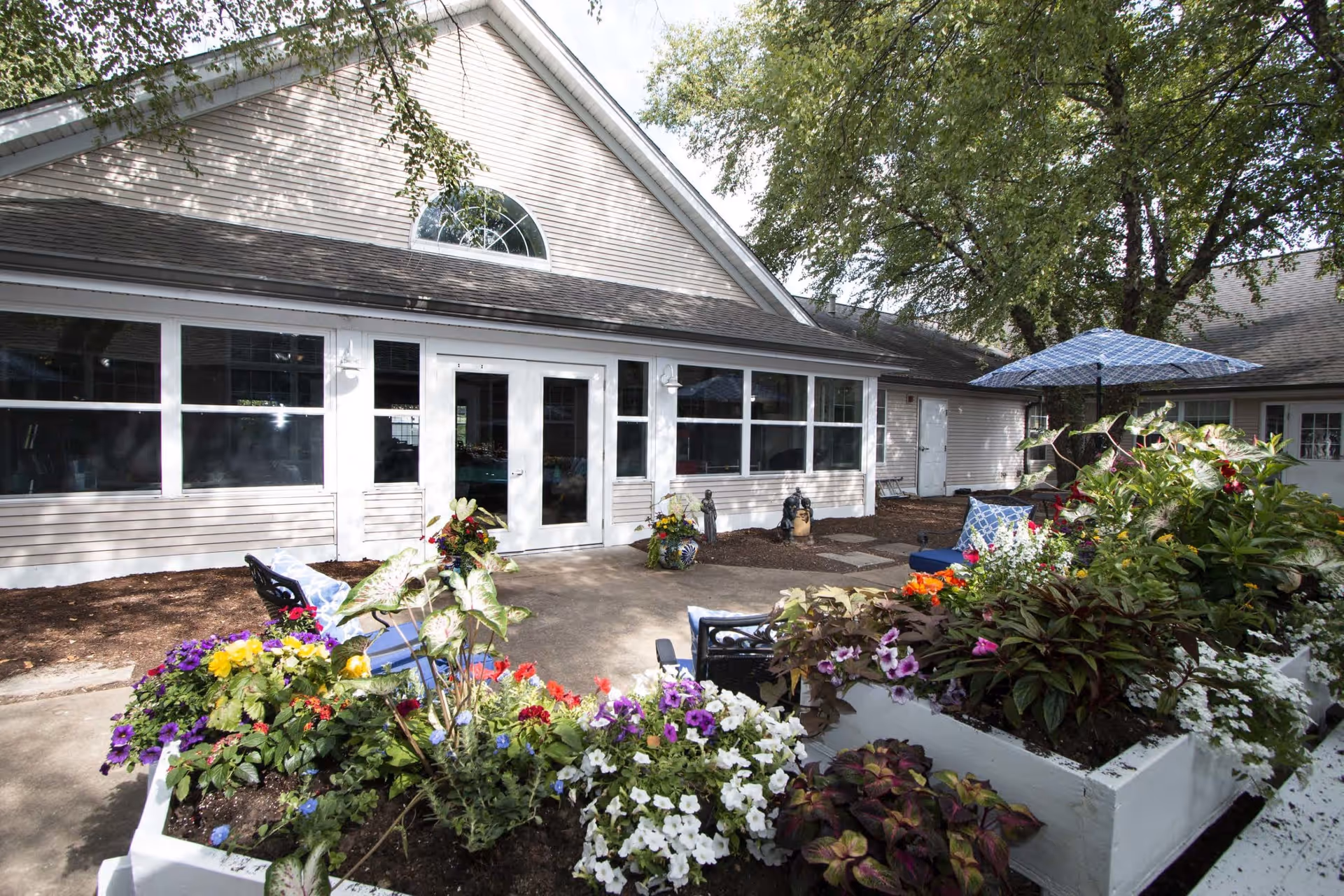 Outdoor patio area at St. Andrew's Assisted Living of Bridgeton featuring a concrete walkway, colorful flower beds, patio chairs with cushions, and a blue umbrella. The building exterior is light-colored with large windows and a peaked roof. Trees provide shade over the seating area.