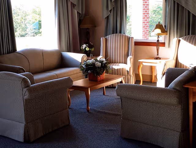 A cozy living room area with a beige sofa, two beige armchairs with striped upholstery, a wooden coffee table with a floral arrangement, and a wooden side table with a lamp. Large windows with curtains allow natural light to brighten the room.