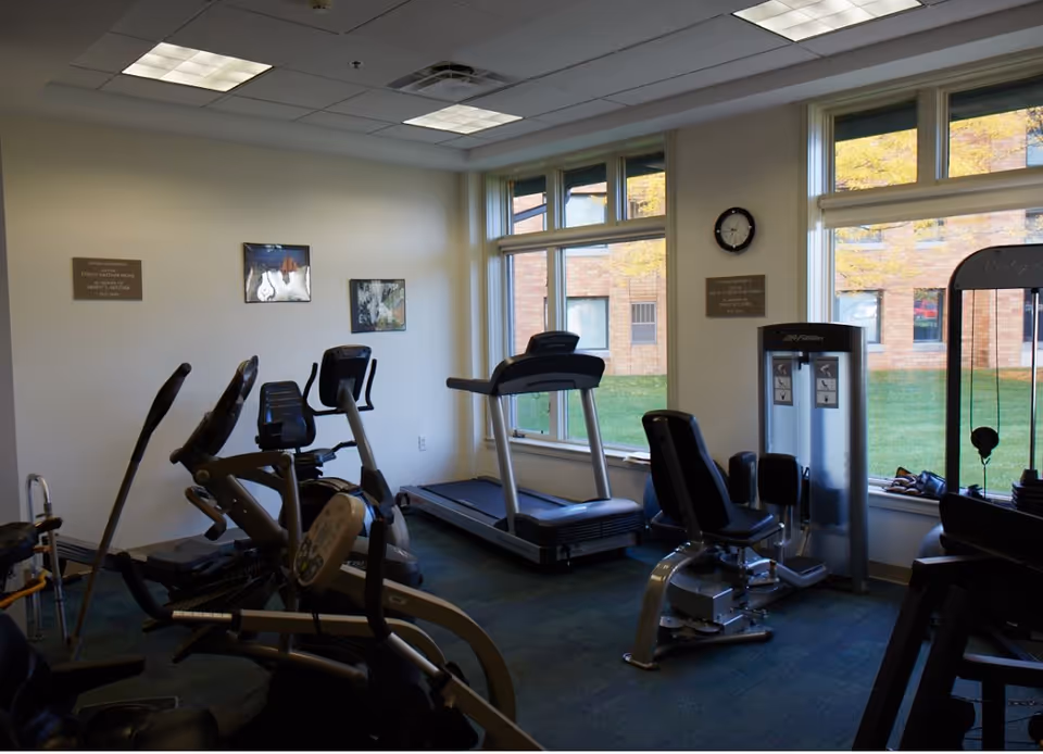 A small fitness room with exercise equipment including a treadmill, stationary bikes, and weight machines. The room has large windows showing a grassy outdoor area and a brick building outside. There are plaques and a clock on the walls.