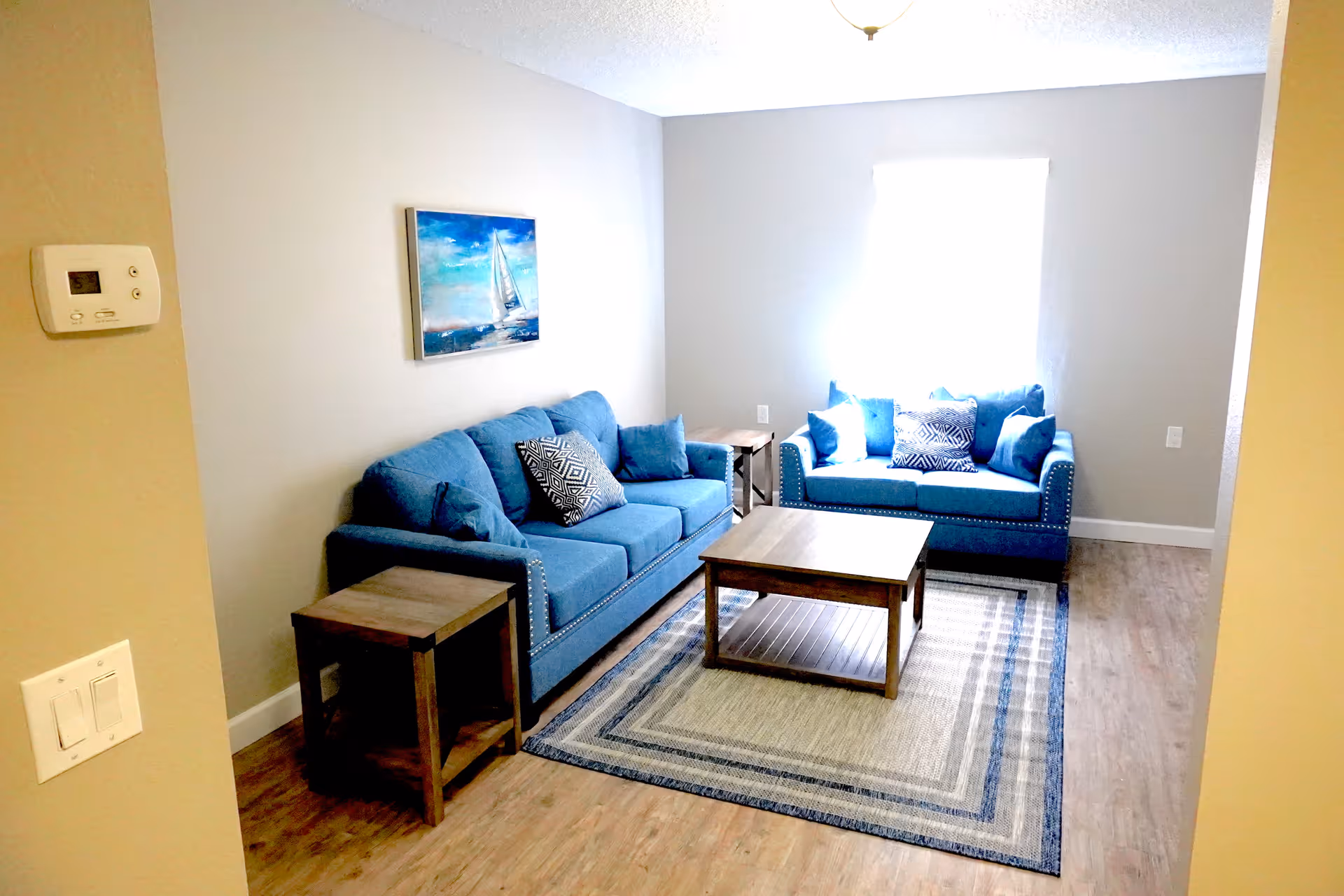 A small living room with two blue sofas adorned with patterned and solid blue cushions. There is a wooden coffee table on a beige and blue area rug, flanked by two matching wooden side tables. A painting of a sailboat on the water hangs on the wall above one sofa. The room has light-colored walls, a window with bright light coming through, and wood flooring.