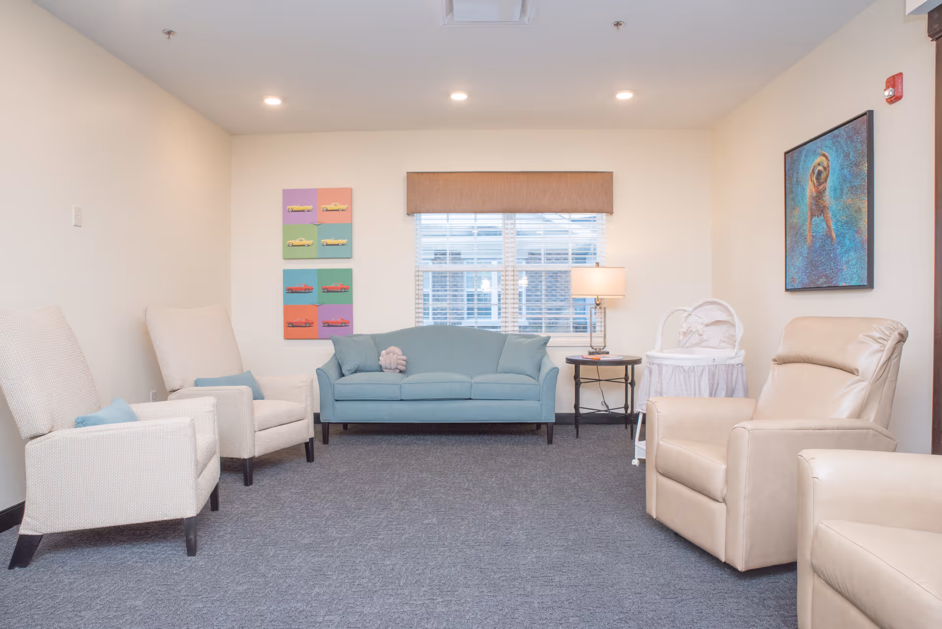A cozy living room with a light blue sofa, two beige armchairs, and a beige recliner. There is a window with blinds and a brown valance in the center of the back wall. A small round side table with a lamp is next to the sofa. Colorful artwork featuring cars is on the left wall, and a painting of a dog is on the right wall. A white bassinet is also visible near the recliner.