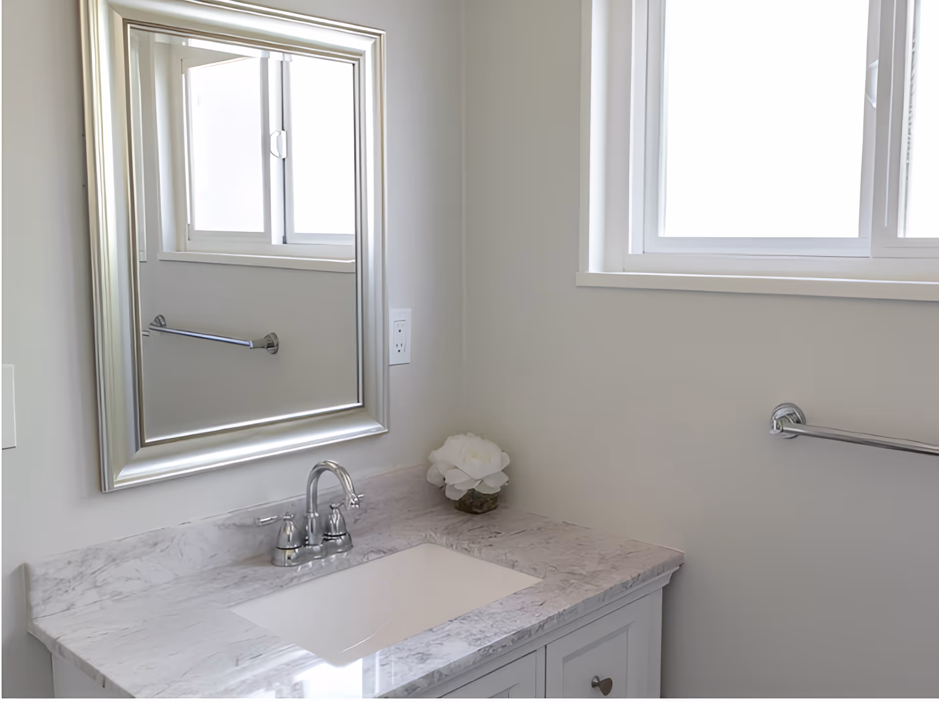 A clean and modern bathroom sink area with a marble countertop, a silver framed mirror above the sink, a chrome faucet, a white flower decoration in a small vase, a towel bar on the wall, and a window letting in natural light.