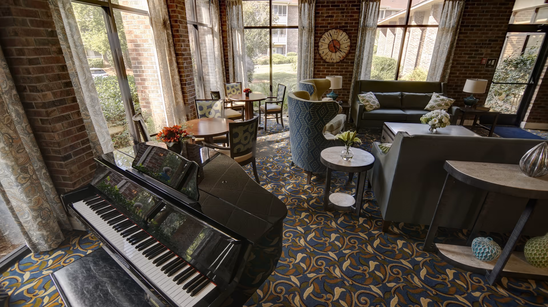 A cozy living room area with a black grand piano in the foreground, patterned carpet, several round tables with chairs, a green sofa, armchairs, side tables with lamps and flowers, large windows with sheer curtains letting in natural light, and a decorative wall clock on a brick wall.