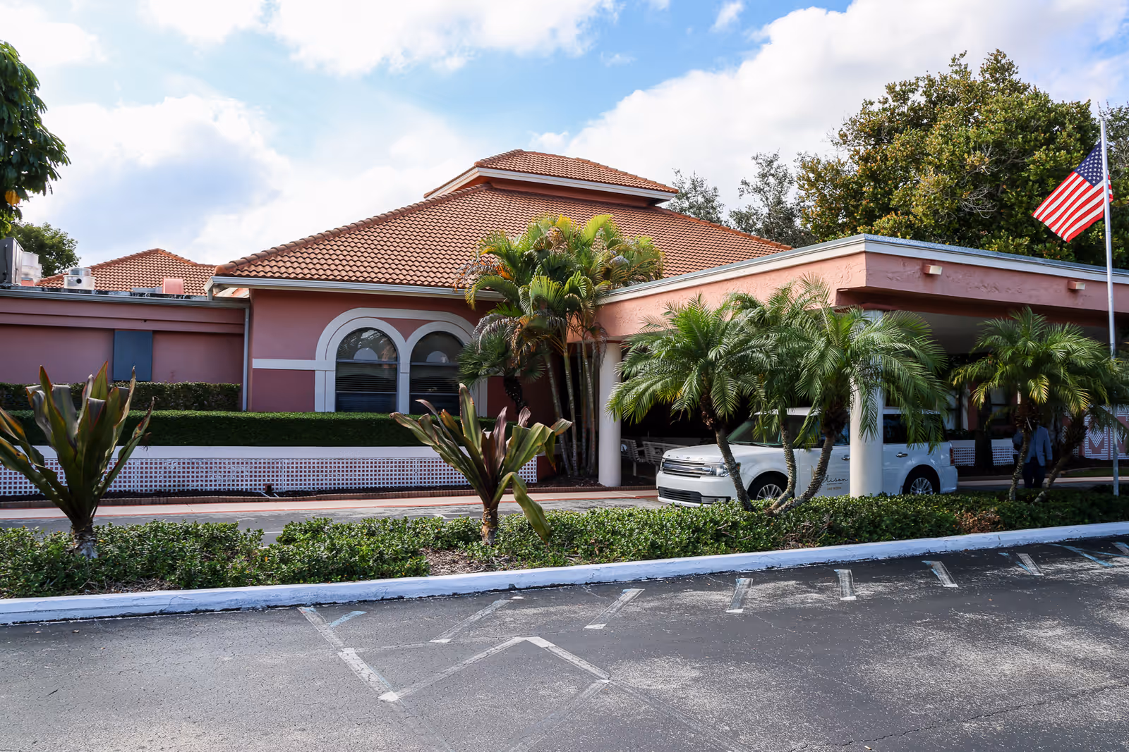 Exterior view of a single-story building with a terracotta tiled roof and pink walls, surrounded by palm trees and greenery. There is a white vehicle parked under a covered entrance, and an American flag is visible on a flagpole to the right. The sky is partly cloudy.