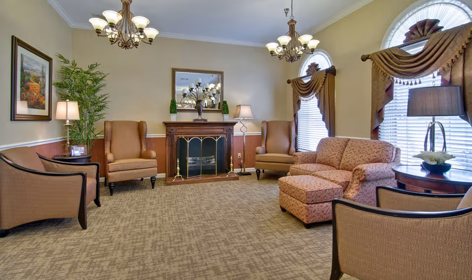 A cozy living room with beige walls and carpet, featuring a fireplace with a decorative screen and a mirror above it. The room has two large armchairs, a patterned sofa with an ottoman, two side tables with lamps, a potted plant, framed artwork, and two windows with ornate drapes.