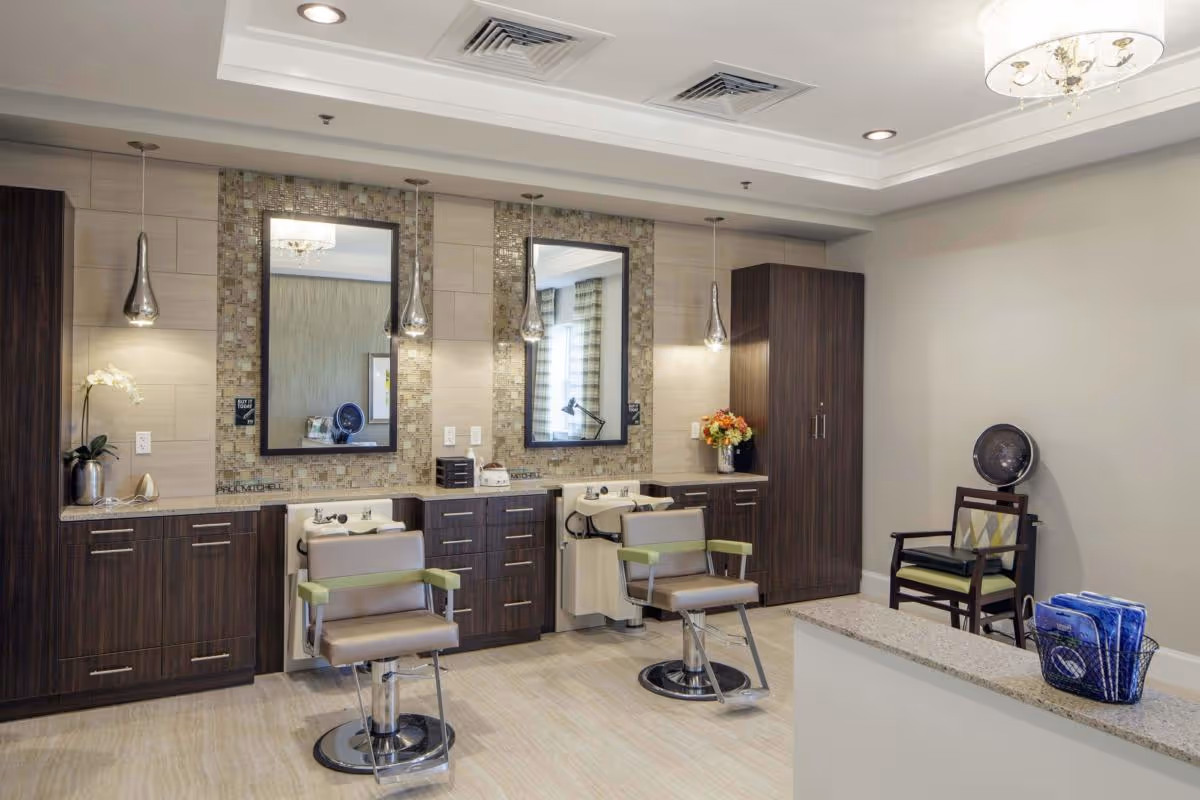 Interior view of a salon area in a senior living facility featuring two salon chairs in front of two large mirrors with mosaic tile backsplashes. The space includes dark wood cabinetry, pendant lights, a small waiting area with a chair and a hair dryer, and a countertop with a basket of blue towels.