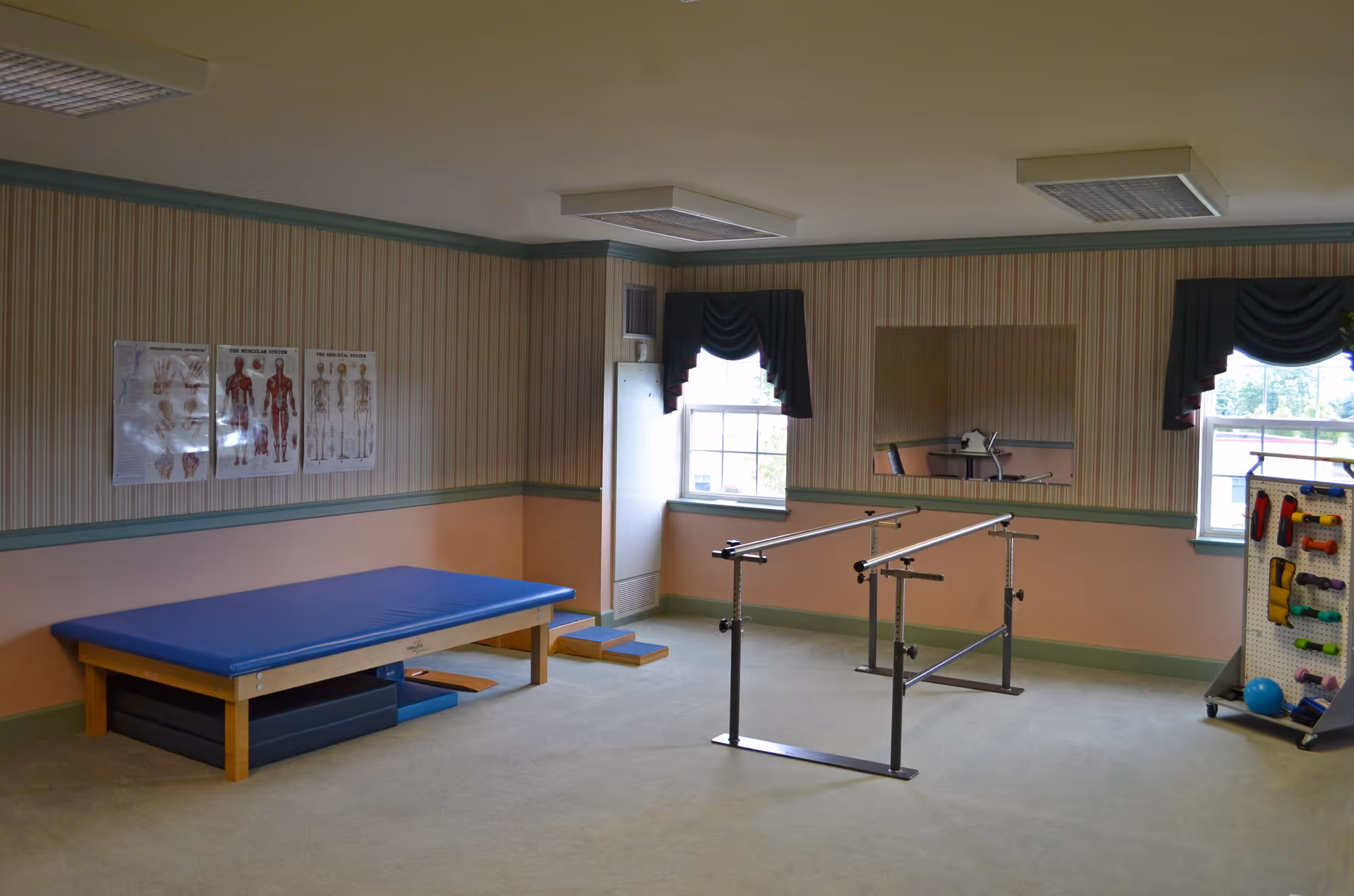 A physical therapy room with a blue padded treatment table, parallel bars for walking exercises, anatomical charts on the wall, two windows with dark green valances, a large mirror, and a rack holding various therapy equipment and exercise balls.
