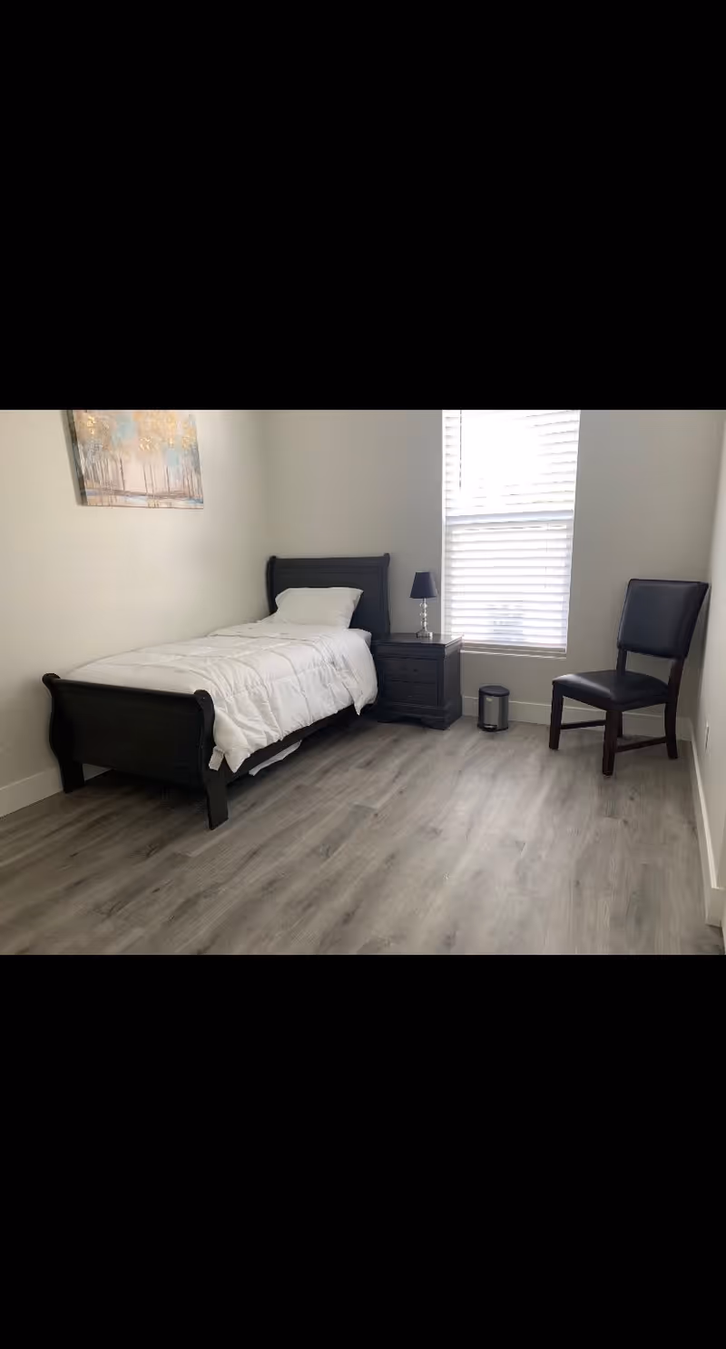 A simple bedroom with a single black wooden bed with white bedding, a black nightstand with a small lamp, a black chair, a trash bin, and a window with blinds letting in natural light. There is a painting on the wall above the bed.