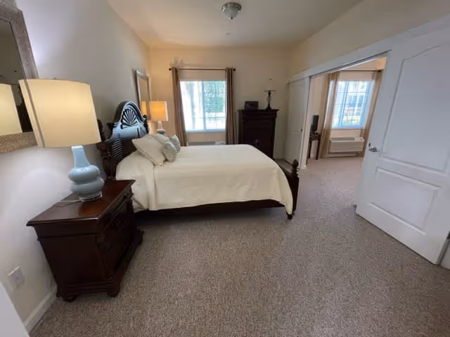 A bedroom with a large bed covered in a white quilt and several pillows. There are two wooden nightstands on either side of the bed, each with a lamp. A window with brown curtains is visible on the far wall, and a wooden dresser with a lamp and a small TV is against the right wall. An open door leads to an adjacent room with a window and an air conditioning unit.