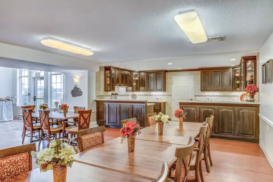 Bright communal dining room with long wooden tables, floral centerpieces, chairs, and a kitchenette/bar with dark wood cabinets.