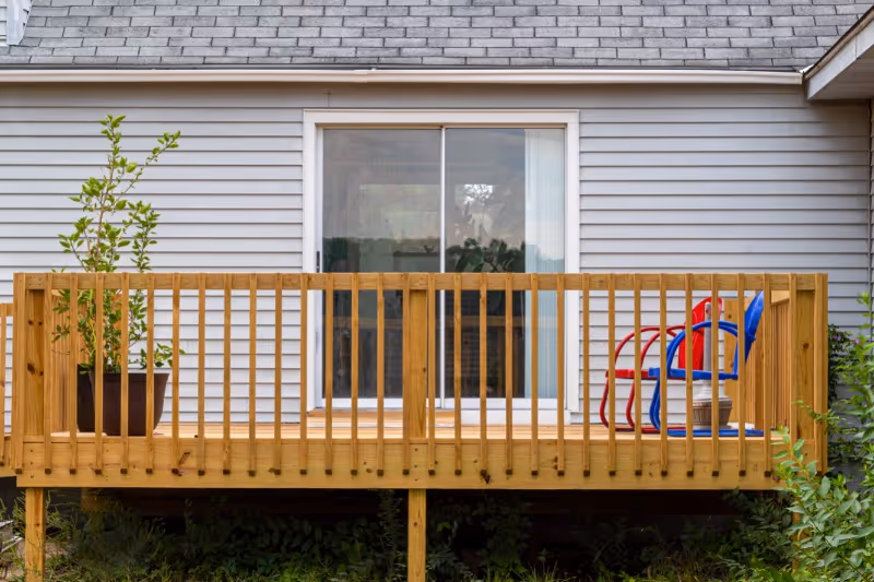 A wooden deck attached to a house with gray siding and a sliding glass door. The deck has a wooden railing, a potted plant on the left side, and two colorful chairs, one red and one blue, on the right side.