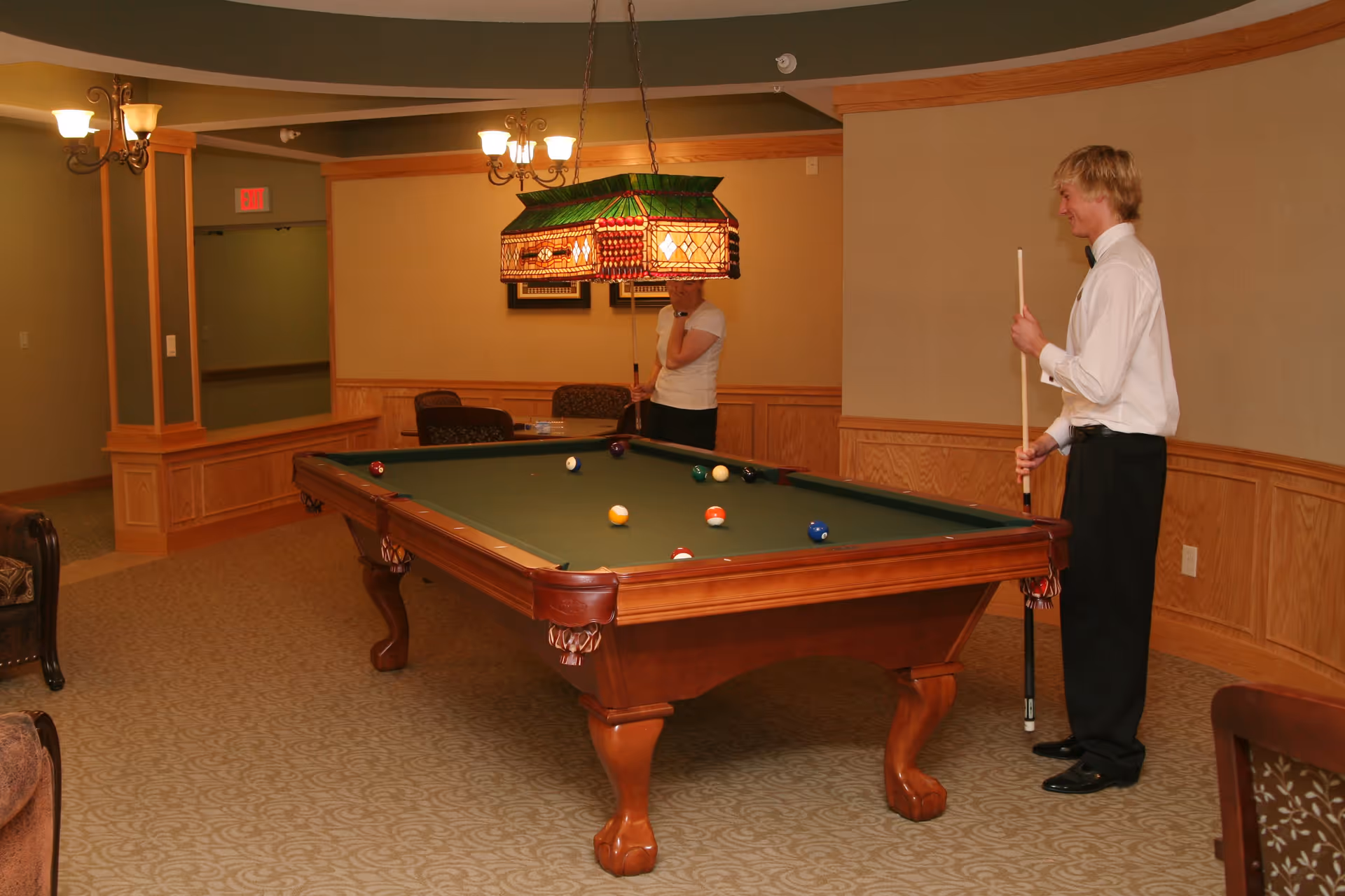 Two people playing pool in a room with beige walls and carpet. The room has wooden paneling along the lower walls and a green pool table with balls arranged on it. There is a stained glass hanging light fixture above the pool table and some chairs and tables in the background.