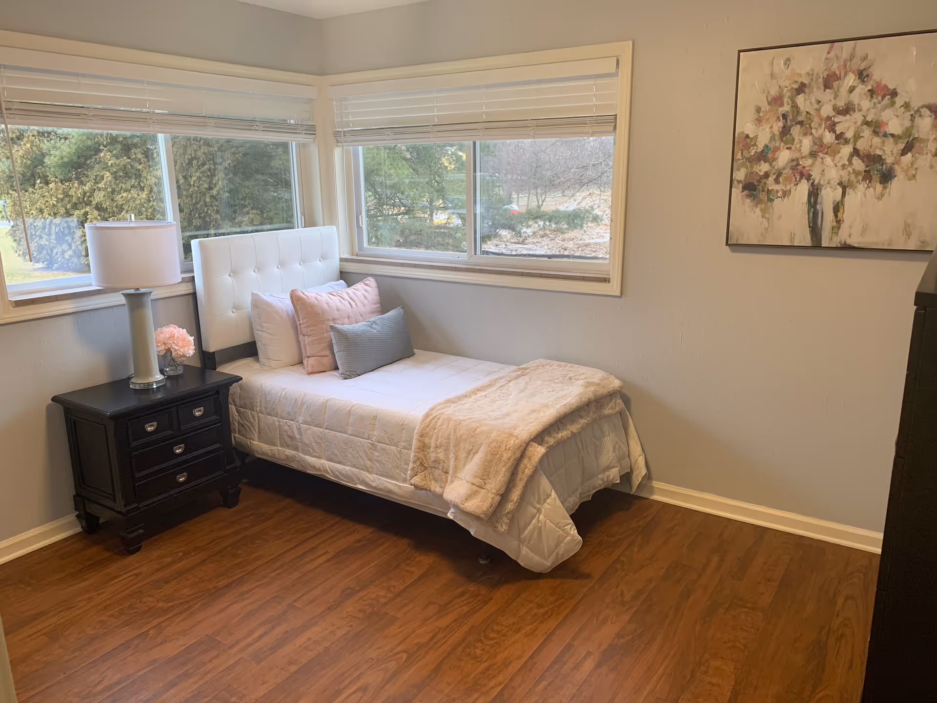 A small bedroom with a single bed featuring a white tufted headboard, white bedding, and several pillows in white, pink, and gray. Next to the bed is a black nightstand with three drawers, a white lamp, and a small pink flower arrangement. The room has wooden flooring, two large windows with white blinds, and a floral painting on the wall.