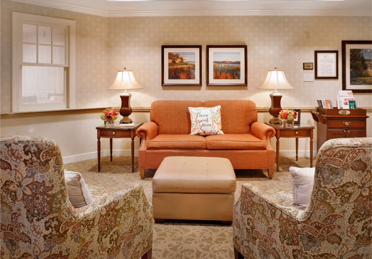 A cozy living room area with an orange sofa featuring a decorative pillow that says 'Home Sweet Home'. Two patterned armchairs face the sofa with a beige ottoman in between. There are two wooden side tables with lamps and flower arrangements on either side of the sofa. The walls have framed landscape artwork and a window on the left side.