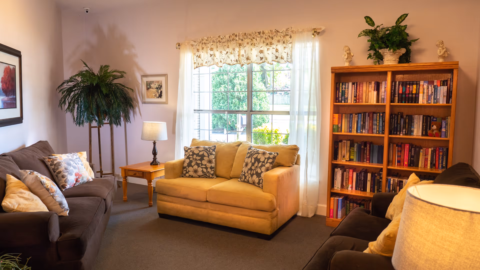 A cozy living room in Southern Care Assisted Living featuring two dark brown sofas and one beige loveseat with patterned cushions. There is a wooden side table with a lamp between the sofas, a tall green plant in the corner, a large window with sheer white curtains letting in natural light, and a wooden bookshelf filled with books and decorative items.